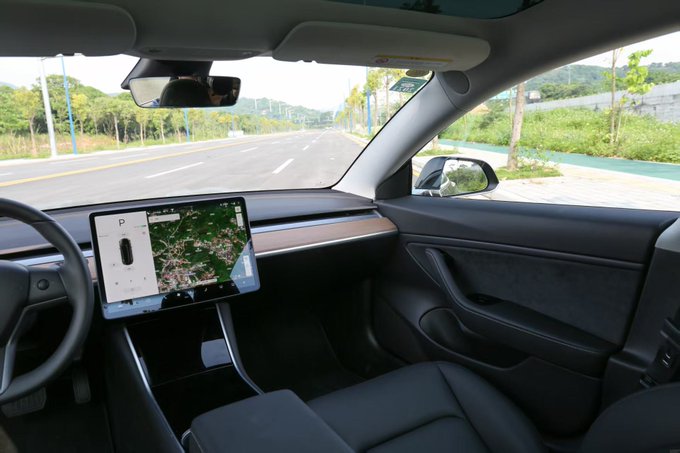 Interior view from the passenger seat of a Tesla, showing the central touchscreen displaying a map in Park mode and a view of an empty street through the windshield.