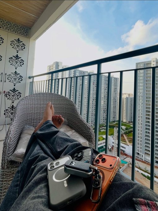 A first-person view of someone relaxing on a high-rise balcony, with three smartphones and AirPods resting on a blanket over their lap.