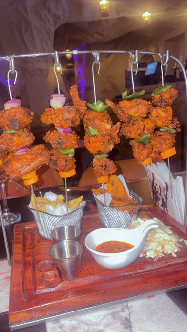 A hanger-style skewer display showcases grilled, marinated meat pieces alternated with green peppers, red onions, and carrots, accompanied by a basket of fries, tortilla chips, and a small metal cup with a bowl of red dipping sauce. The dish appears to be a hearty, restaurant-style kebab platter that humorously substitutes for a simpler meal in the caption's playful reference.