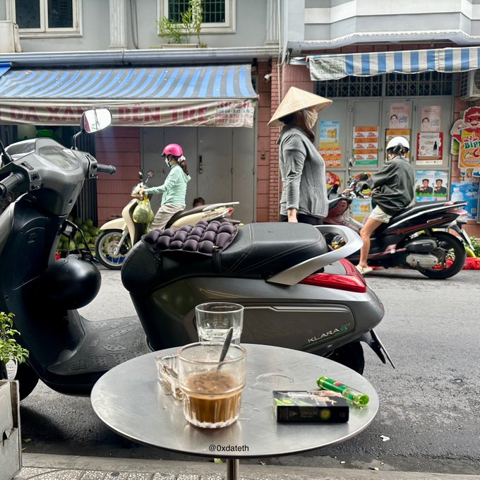 A street scene taken from a cafe table with coffee and cigarettes, showing a person in a conical hat on a black scooter loaded with purple produce, with other scooters and pedestrians on the street.