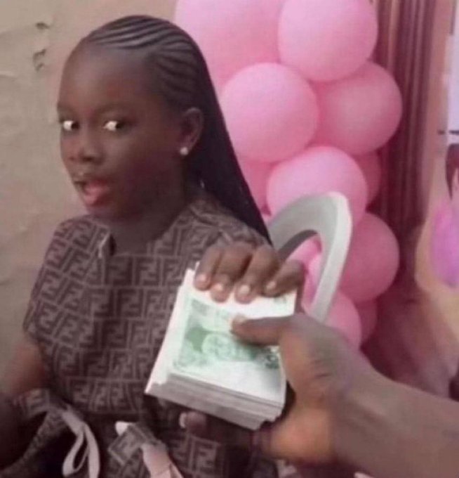 A young girl with braided hair wearing a patterned brown outfit holds a stack of US dollar bills, looking surprised, with pink balloons in the background as another hand reaches toward the money.