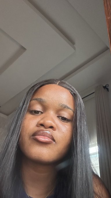 A low-angle close-up selfie of a woman with long straight dark hair and glossy lips, taken indoors with a white paneled ceiling visible above her.