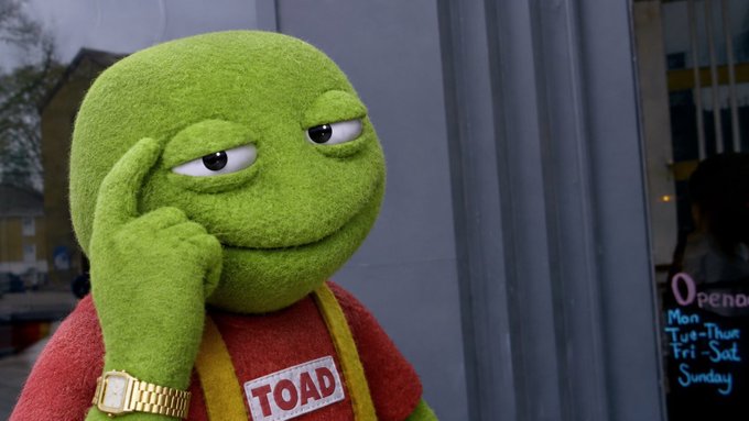 A green frog-like puppet character (labeled 'TOAD') wearing a red shirt and gold watch, touching its temple thoughtfully while standing outside a storefront.