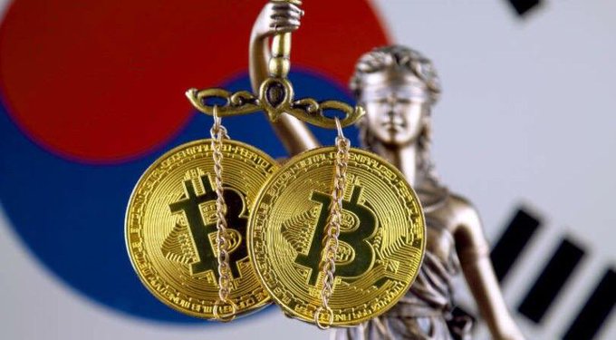 Gold Bitcoin tokens hanging from chains held by a Lady Justice statue, with the South Korean flag (Taegukgi) blurred in the background.