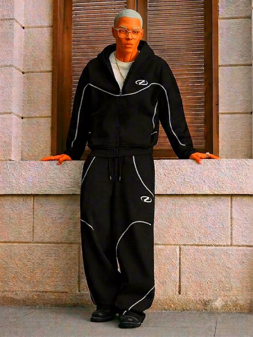 A man wearing glasses models a black tracksuit with white piping and a small logo, posing against a stone wall.