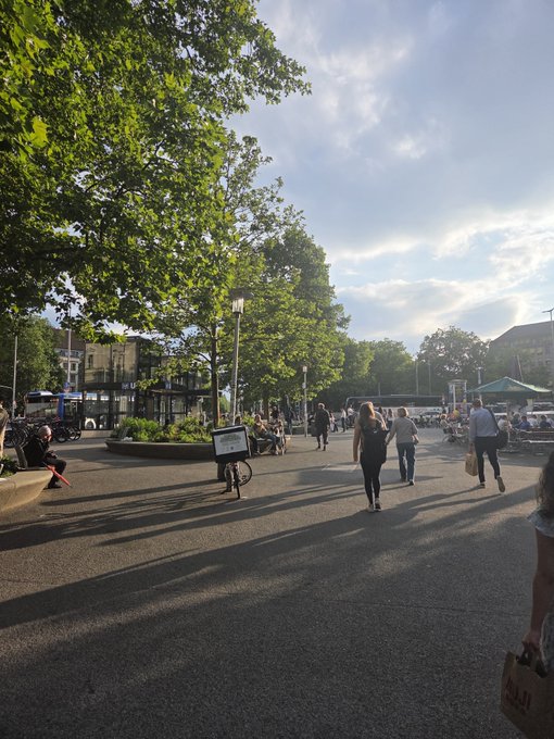 A sunny outdoor plaza scene with large leafy trees, pedestrians walking around, and people seated on benches and at outdoor café tables.