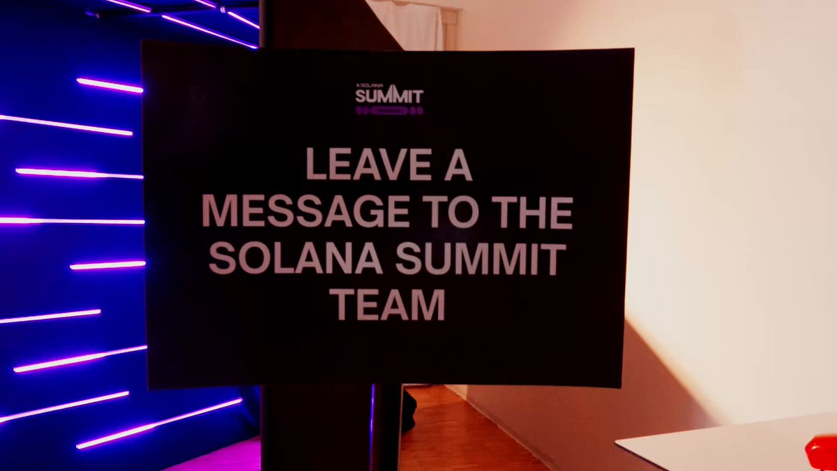 The image features a black sign with white text that reads "LEAVE A MESSAGE TO THE SOLANA SUMMIT TEAM". The sign is placed near a purple light strip, which adds a vibrant touch to the scene. In the background, there are two people standing next to each other and a red ball on the floor.