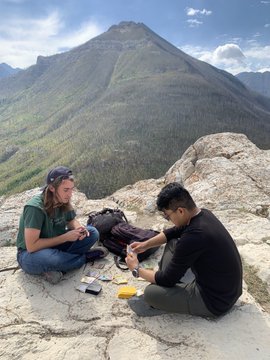 Two people sitting on a rocky mountain summit with a scenic view of forested peaks and valleys, appearing to share a meal together with a yellow container visible between them.