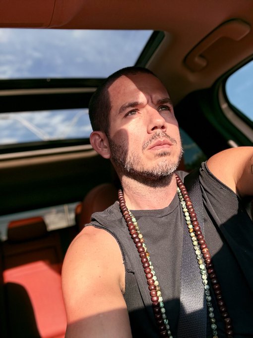 A photograph of a man with a short beard wearing a gray tank top and beaded necklaces, sitting inside a vehicle with a sunroof open to the sky.