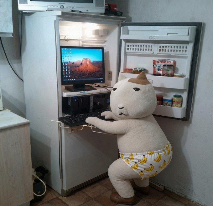 A humorous image of a chubby cat figurine wearing banana-print underwear, standing at a refrigerator that has been converted into a computer setup with keyboard, mouse, and monitor.