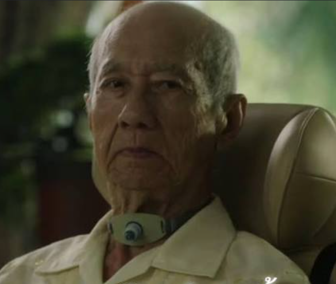 An elderly bald man wearing a neck brace sits in a chair, looking off-camera with a serious expression.