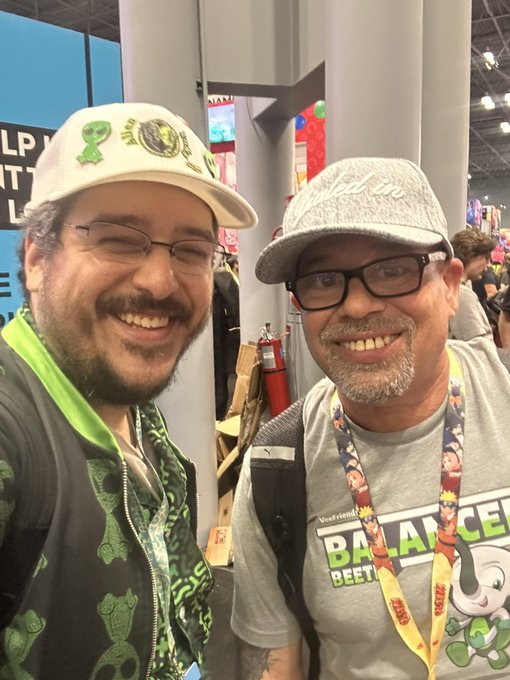 Two smiling men pose together at what appears to be a convention, one wearing a white cap with green emblems and a green hoodie, the other in a gray 'Balance' t-shirt and cap.