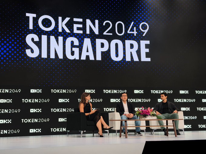 A panel discussion at TOKEN2049 Singapore features a moderator interviewing two guests on stage, with a large "TOKEN 2049 SINGAPORE" backdrop and OKX sponsor logos, tied to the announcement of Samsung bringing stablecoins to 82 million Galaxy devices in partnership with Solana.