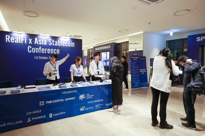 A photo of a conference entrance area with a large blue sign reading 'RealFi x Asia Stablecoin Conference 1st October 2024,' showing staff at a registration desk with logos for 'THE CAPITAL SUMMIT,' 'ASIA STABLECOIN ALLIANCE,' 'SB DigiTrust,' 'Topfinance,' 'PHAROS,' and 'LayerZero,' while attendees interact in the foreground.