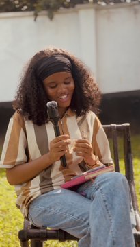 A young woman with curly hair and a striped shirt sits outdoors holding a microphone and smiling.