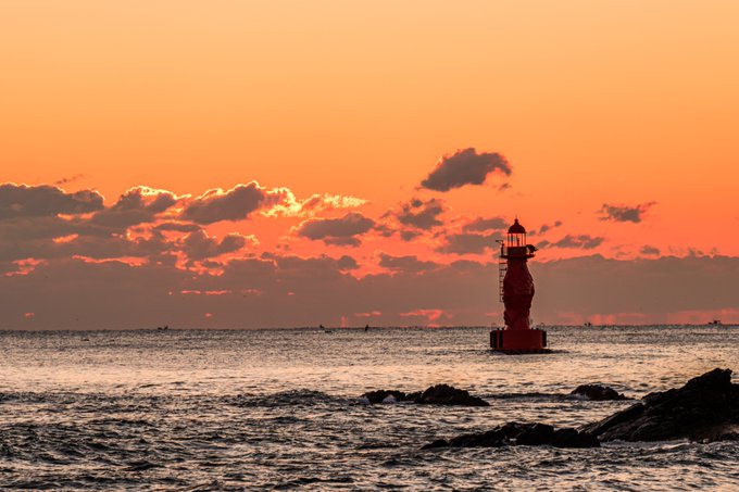 A scenic sunset view over the ocean features a red lighthouse silhouetted against a bright orange sky with scattered clouds.