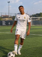 A photo of a young soccer player wearing a white Real Madrid 'Emirates Fly Better' jersey (number 7) standing on a grass field with a soccer ball at his feet.
