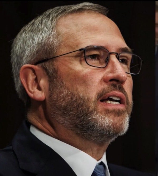 A close-up photograph of an older man with a gray beard, wearing glasses and a dark suit with a tie, appearing to be speaking.