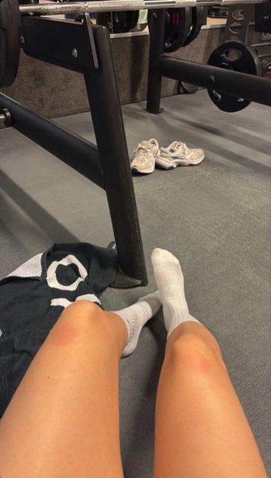 First-person view of someone's legs extended on a gym floor in white socks, with a pair of white sneakers visible near weight equipment in the background.