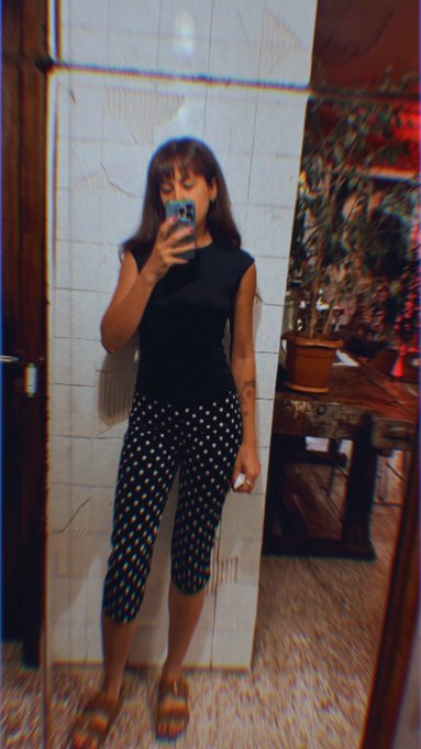 A woman takes a mirror selfie in a black sleeveless top and black polka-dot capri pants, standing in front of a tiled wall.