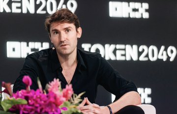 A young man with short brown hair, wearing a dark button-up shirt, speaks at the TOKEN2049 conference with a pink floral arrangement in the foreground, embodying the event's polished, high-energy atmosphere.