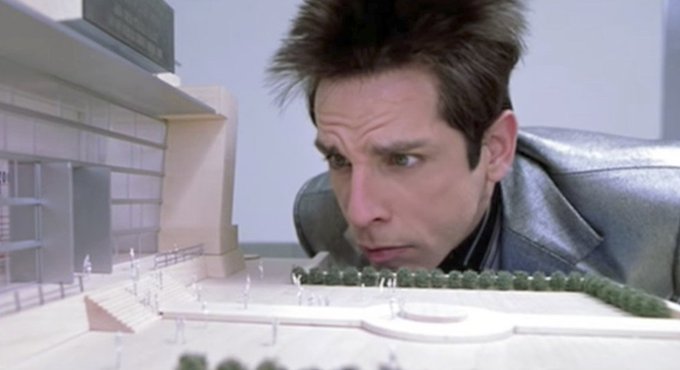 A man with exaggerated spiky hair, resembling the character Zoolander, looking closely at an architectural scale model.