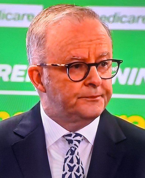 A close-up of Australian Prime Minister Anthony Albanese wearing glasses and a patterned blue tie, standing in front of a green background with the word 'medicare' visible.