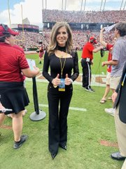 A smiling woman in a black jumpsuit stands on the sidelines of a packed football stadium, seemingly unaffected by the post's claim about anxious crypto traders rushing to their screens. The image's relaxed, upbeat atmosphere contrasts sharply with the implied financial urgency of the caption.