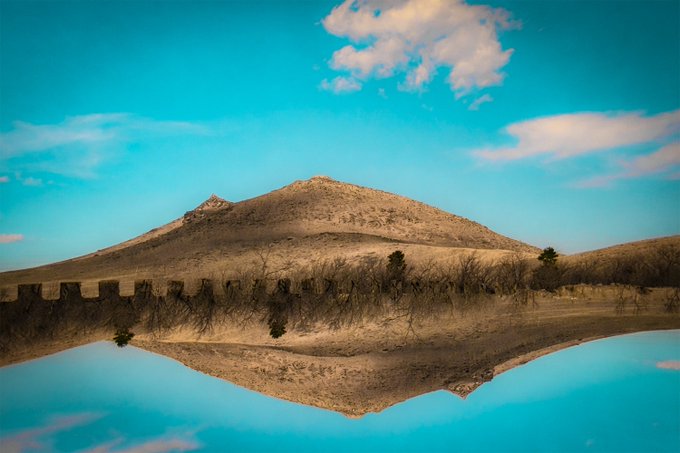 A serene landscape featuring a mountain and crenellated wall perfectly reflected in calm blue water beneath a sky with scattered white clouds.