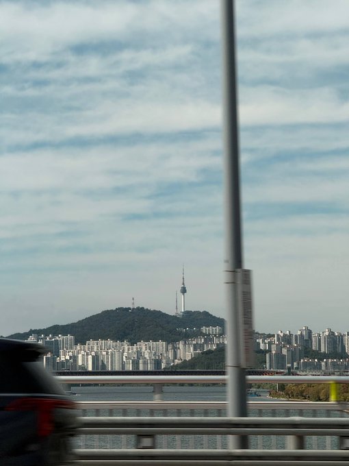 A landscape photograph taken from a moving vehicle showing Seoul's Namsan Tower in the distance, with the Han River, city skyline, and a bridge railing in the foreground.