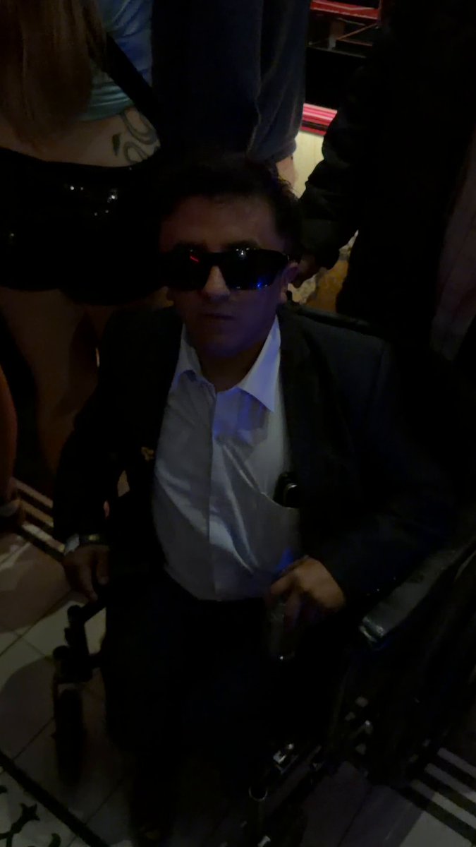 The image shows a man wearing sunglasses, sitting in a wheelchair with his arms resting on the backrest of the chair. The man appears to be dressed in formal attire, possibly for an event or gathering. There are other people present in the scene as well, suggesting that this might be a social event or gathering where attendees are seated and engaged in conversation.