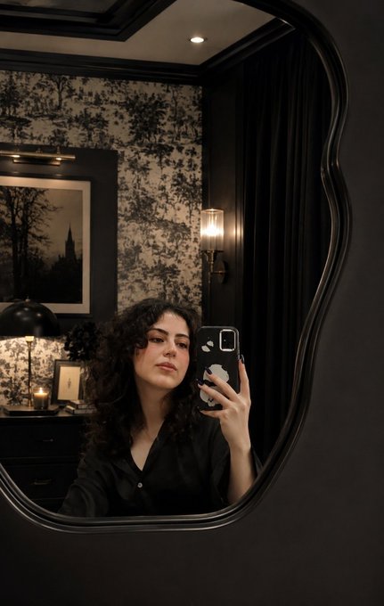 The image features a woman taking a selfie with her cell phone, holding it up against a mirror that has an interesting shape resembling a window or a door frame. The background of the photo includes a black wall adorned with wallpaper featuring trees, which adds a touch of nature and elegance to the scene. The woman is dressed in a black outfit, and there are two candles on a dresser nearby, adding a cozy ambiance to the room.