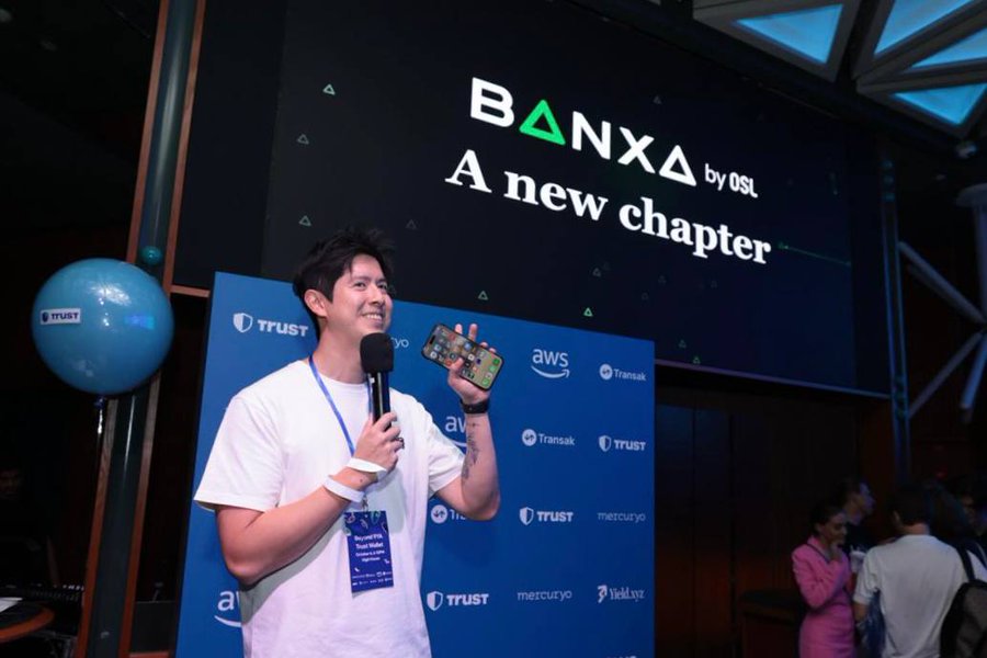 A man with a name tag stands on stage at a Trust Wallet and Token2049 event, holding a microphone and phone in front of a large screen displaying "BANXA by OSL - A new chapter" alongside sponsor logos including AWS, Trust, Transak, and Metcuryo.