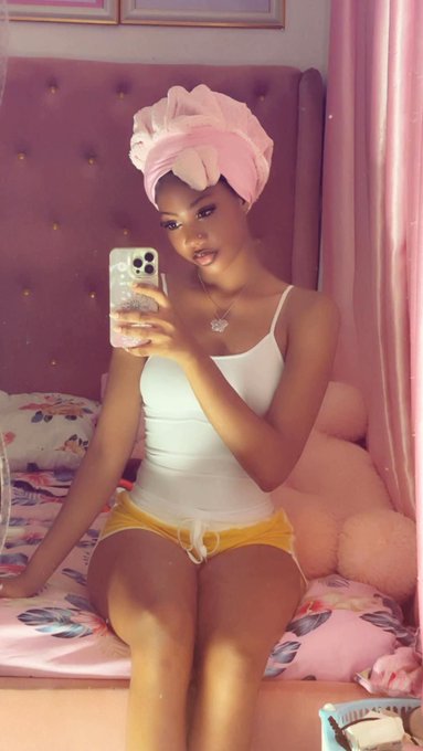A woman wearing a pink towel on her hair sits on top of a bed taking a selfie with a cell phone.