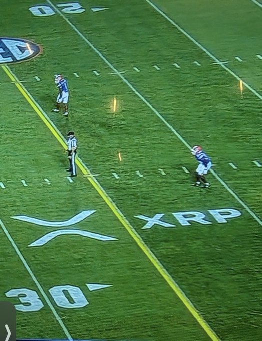 A football game scene shows players on a green field with 'XRP' branding visible near the 30-yard line, alongside a large X logo and SEC conference marking.