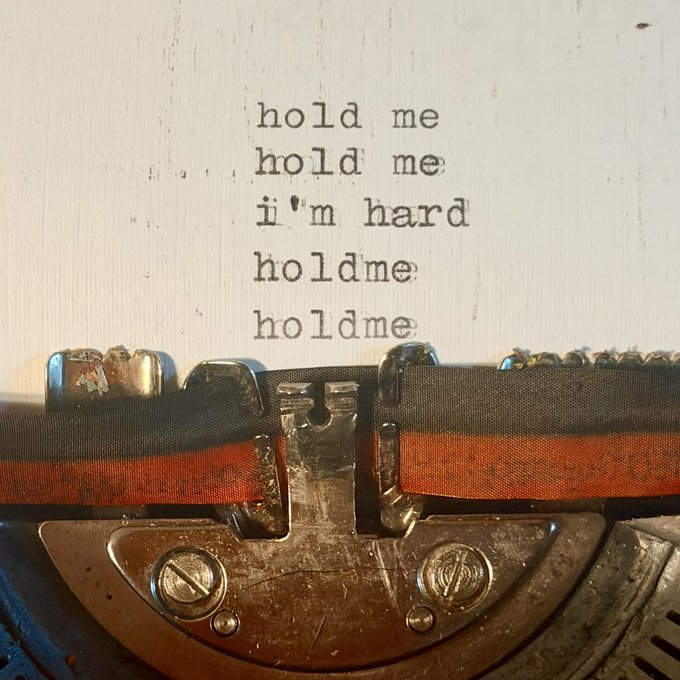 A close-up of a vintage typewriter typing the repeated phrase 'hold me / hold me / i'm hard / holdme / holdme' onto white paper with a red ribbon visible.