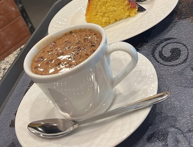 A cup of frothy hot chocolate or coffee sits on a white saucer with a silver spoon, accompanied by a slice of cake on a plate beside it on a textured table.