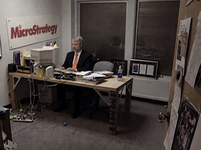 urns of water and a plant sit on the windowsill, with a computer monitor displaying "MicroStrategy" and a man in an orange suit seated at a desk.