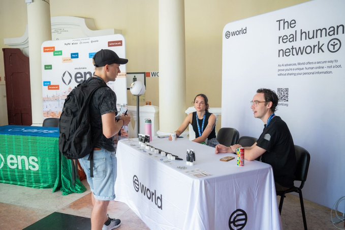 Two booth representatives sitting at a table promoting 'world' as 'the real human network', with a cap-wearing attendee standing in front.