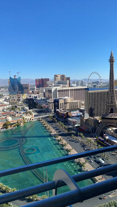 A high-altitude view of the Las Vegas Strip showcases its iconic landmarks, including a replica of the Eiffel Tower, the Bellagio fountains, and the High Roller Ferris wheel under a clear blue sky. The "Gm" greeting pairs with the bright morning ambiance of the bustling cityscape below.