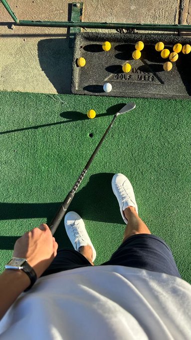 First-person view of someone holding a golf club on artificial turf, with a yellow golf ball positioned to be hit toward a mat labeled 'GOLF MATS' that holds additional balls.