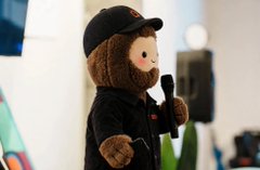 A small brown teddy bear figurine wearing a black cap, black hoodie, and holding a microphone, photographed in what appears to be a gallery setting.