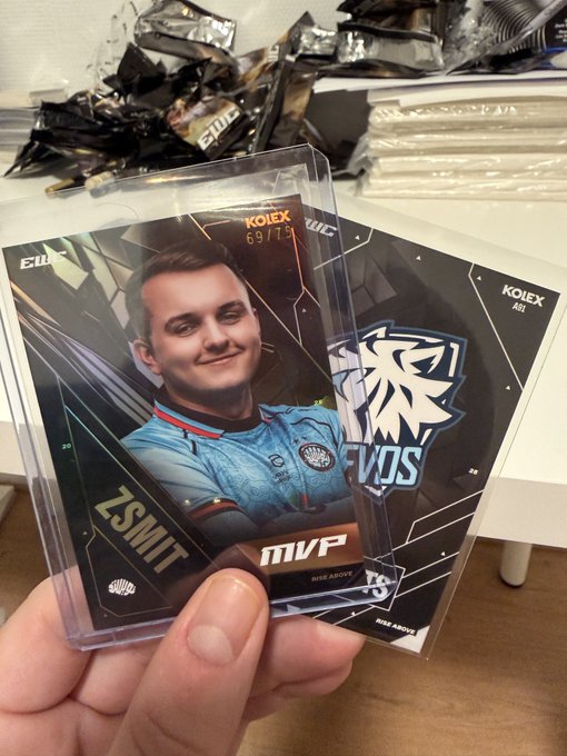 A hand holding two sports trading cards in protective plastic sleeves; the visible card features a soccer player in a light blue jersey, labeled 'KOLEX', 'MVP', and 'ZSMIT'.