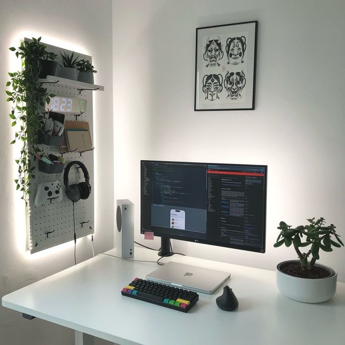A tidy white workspace featuring a monitor displaying code, a mechanical keyboard, a laptop, an Xbox controller, plants, and a framed tiger print on the wall.