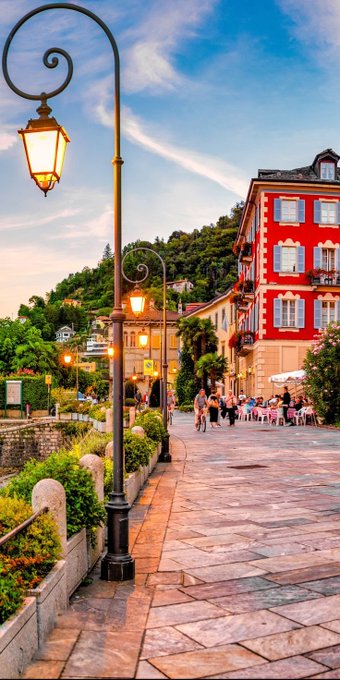 A scenic European street features an ornate black lamppost in the foreground, with a red-brick building, outdoor diners, and lush green hills in the background under a blue sky.