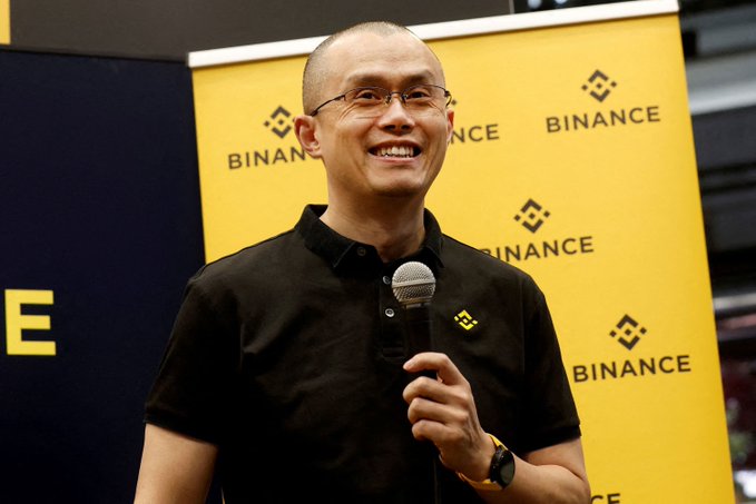 A smiling man wearing a black polo shirt holds a microphone while speaking at an event, standing in front of a yellow backdrop featuring the "BINANCE" logo repeated across it. The image illustrates the accompanying post's claim about CZ (Changpeng Zhao), the former Binance CEO, whose estimated $110 billion net worth now reportedly exceeds that of Bill Gates.
