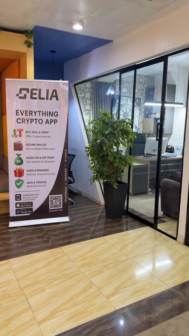 A promotional banner for the "CELIA" Everything Crypto App stands in an office lobby, highlighting features like NFT/Sell & Swap, Secure Wallet, Earn & Rewards, and Safe & Trusted services. The display is positioned next to a potted plant and a glass-walled office space.