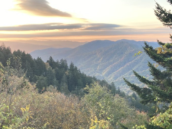 A scenic landscape photograph showing a mountain valley at sunrise or sunset, with forested ridges, autumn-tinged foliage in the foreground, and dramatic clouds in the sky.