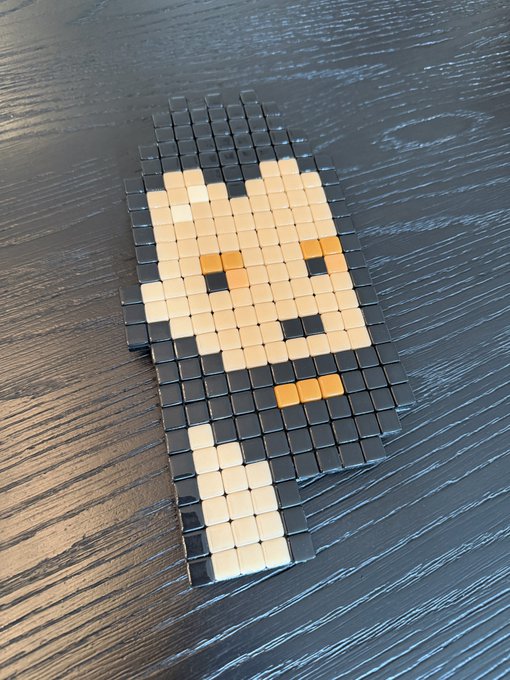 A pixel-art style portrait made of small cube tiles rests on a dark wooden surface, depicting a hooded figure with glowing yellow eyes and a small mouth accent. The physical artwork brings the digital NFT to life, representing the creative pixel-craft pieces made by Ferboccard that were gifted to the recipient.