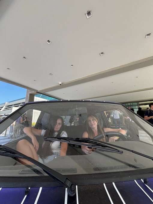 Two young women sitting inside a purple car parked under a covered structure, viewed through the windshield with wipers visible, with other people standing in the background.